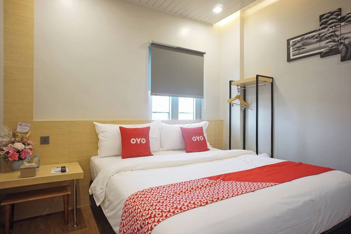 Une chambre d’hôtel comprenant un lit, une table de chevet, une fenêtre avec un store et un porte-manteau fixé au mur.
