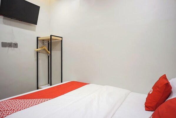 A hotel room with a bed, a red and white bedspread, a red pillow, a TV mounted on the wall, and a coat rack.