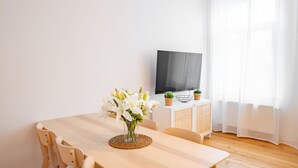 A modern living room with a wooden dining table, a flat-screen TV mounted on a white cabinet, and a large window with sheer curtains.