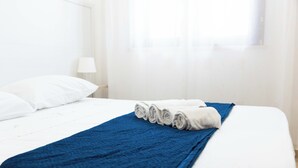 Una cama bien hecha con sábanas blancas y una manta azul, un almohadón blanco y una toalla enrollada al lado.