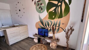 Una sala de estar moderna con piso de madera, un mueble de madera, un televisor sobre un soporte de madera, y una pared con un reloj y un mural de plantas grande.