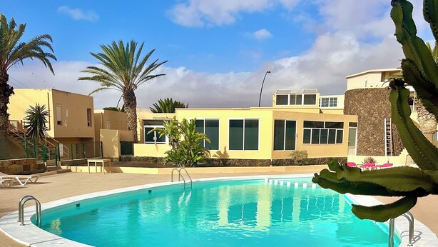 Una piscina con un bordeado de baldosas, palmeras y un edificio con amplias ventanas al fondo.