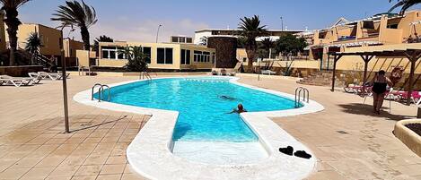 A swimming pool with a tiled deck, surrounded by palm trees and buildings.