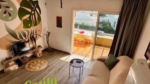 Amplia sala de estar con un sofá grande, una mesita auxiliar y un televisor. Se aprecia una ventana grande con vistas al exterior. La habitación tiene una temática tropical, con una planta grande y un mural en la pared.