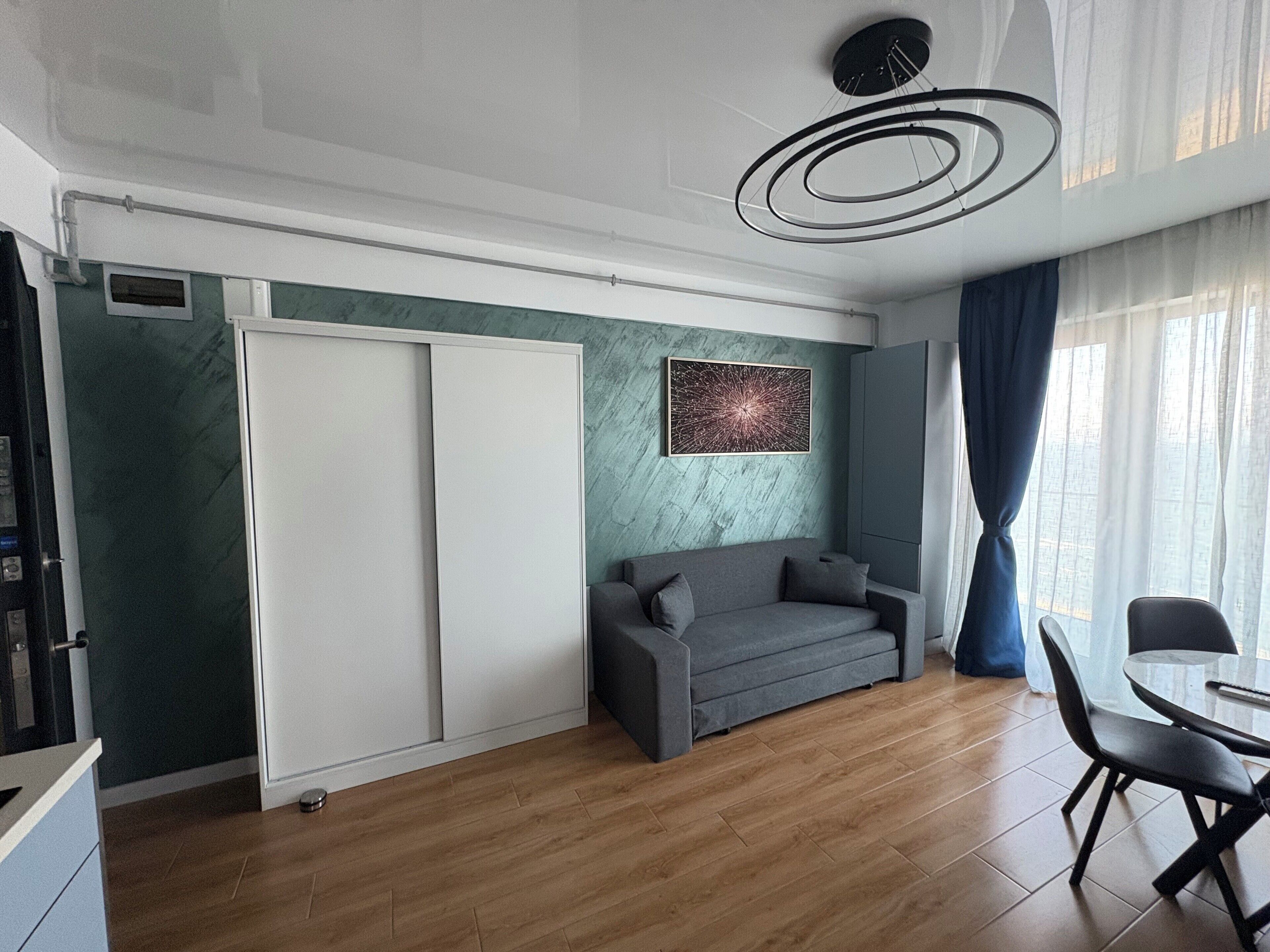 Un salon moderne avec un canapé gris, une table à manger avec des chaises, une grande fenêtre avec des rideaux et un luminaire au plafond.