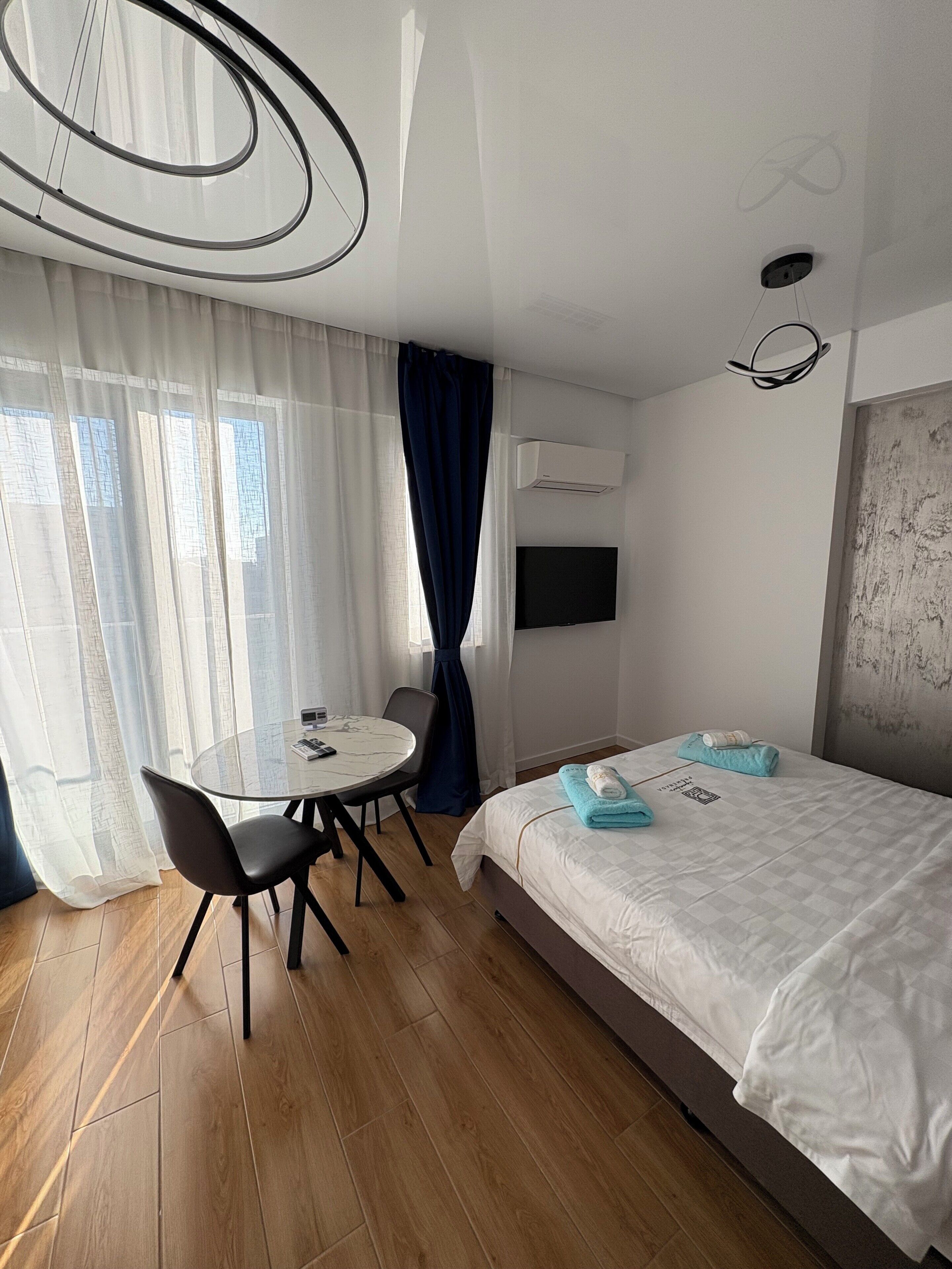 A modern hotel room with a round dining table, a bed with neatly folded towels, and a mounted TV.