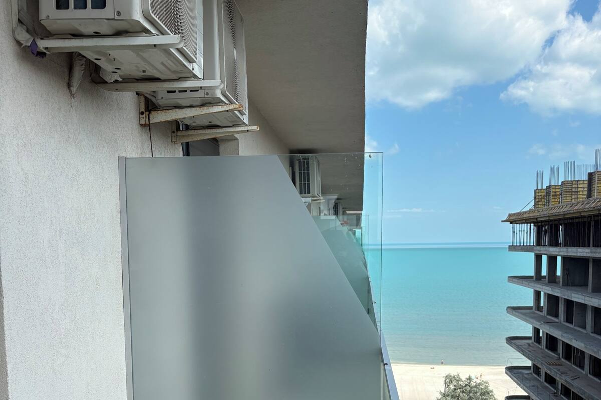 A balcony with a view of the beach and ocean, a glass partition, and a building with multiple floors.