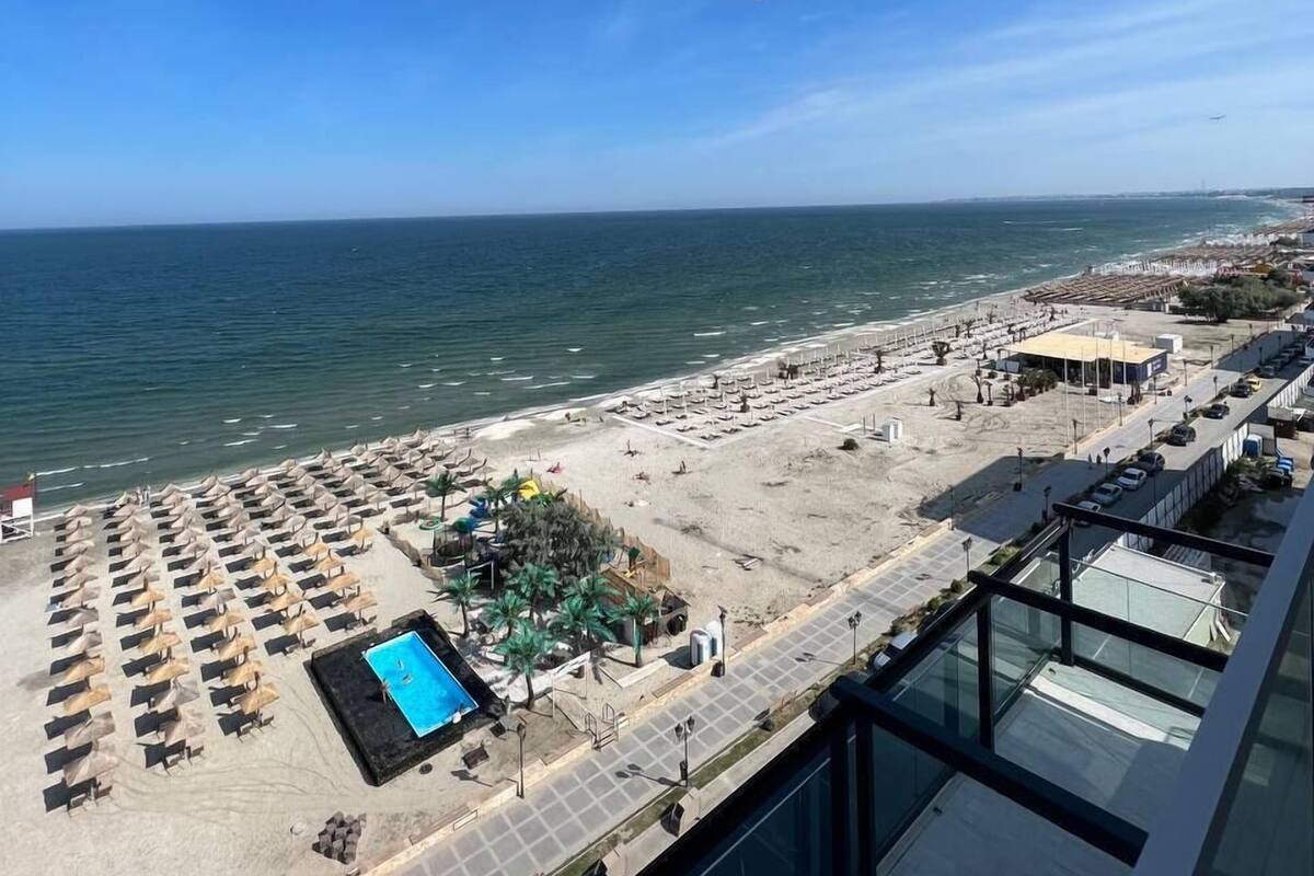 A beach view from a balcony with a pool, lounge chairs, and a parking area.
