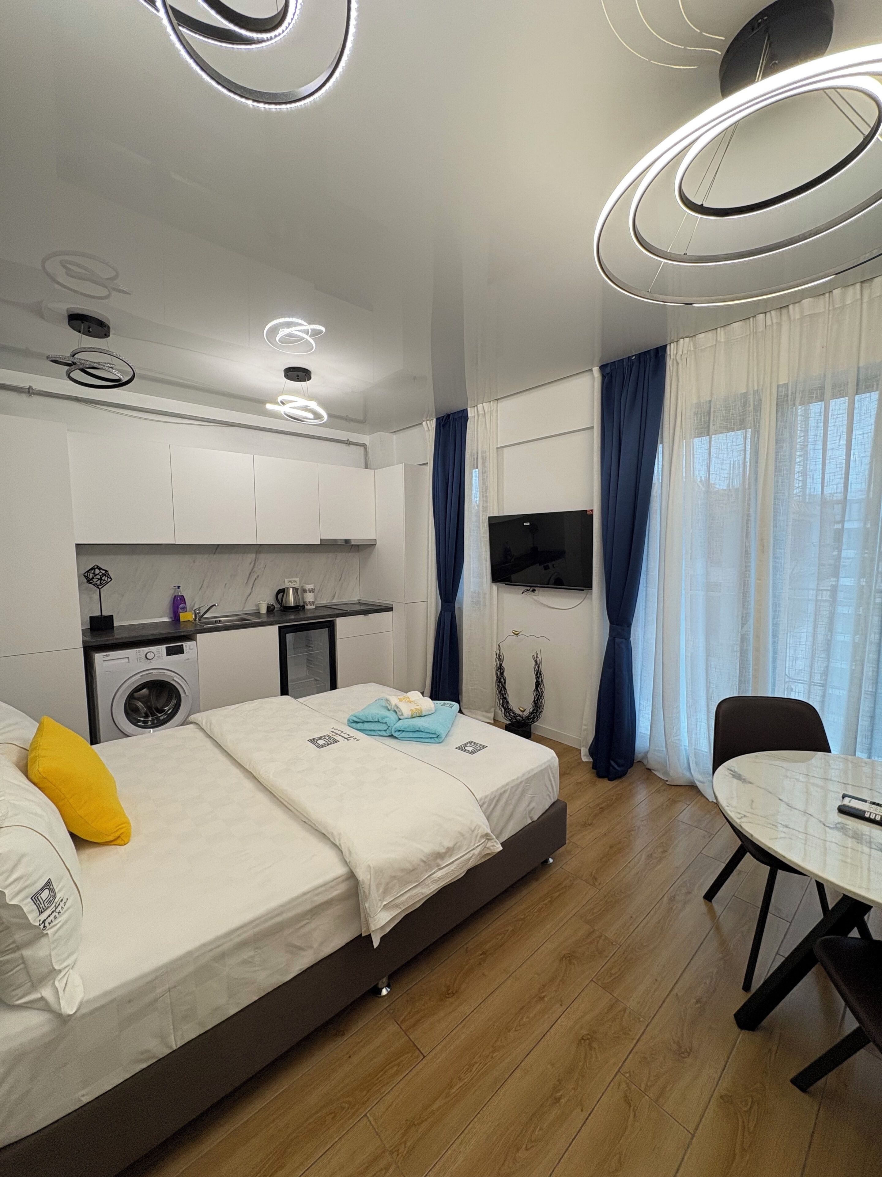 A modern hotel room with a bed, a dining table, a TV, a washing machine, and a kitchen area.