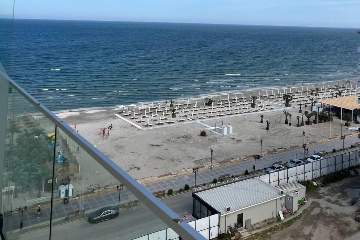A beach view from a high vantage point, featuring a sandy beach with umbrellas and lounge chairs, a parking area, and a clear blue sky.