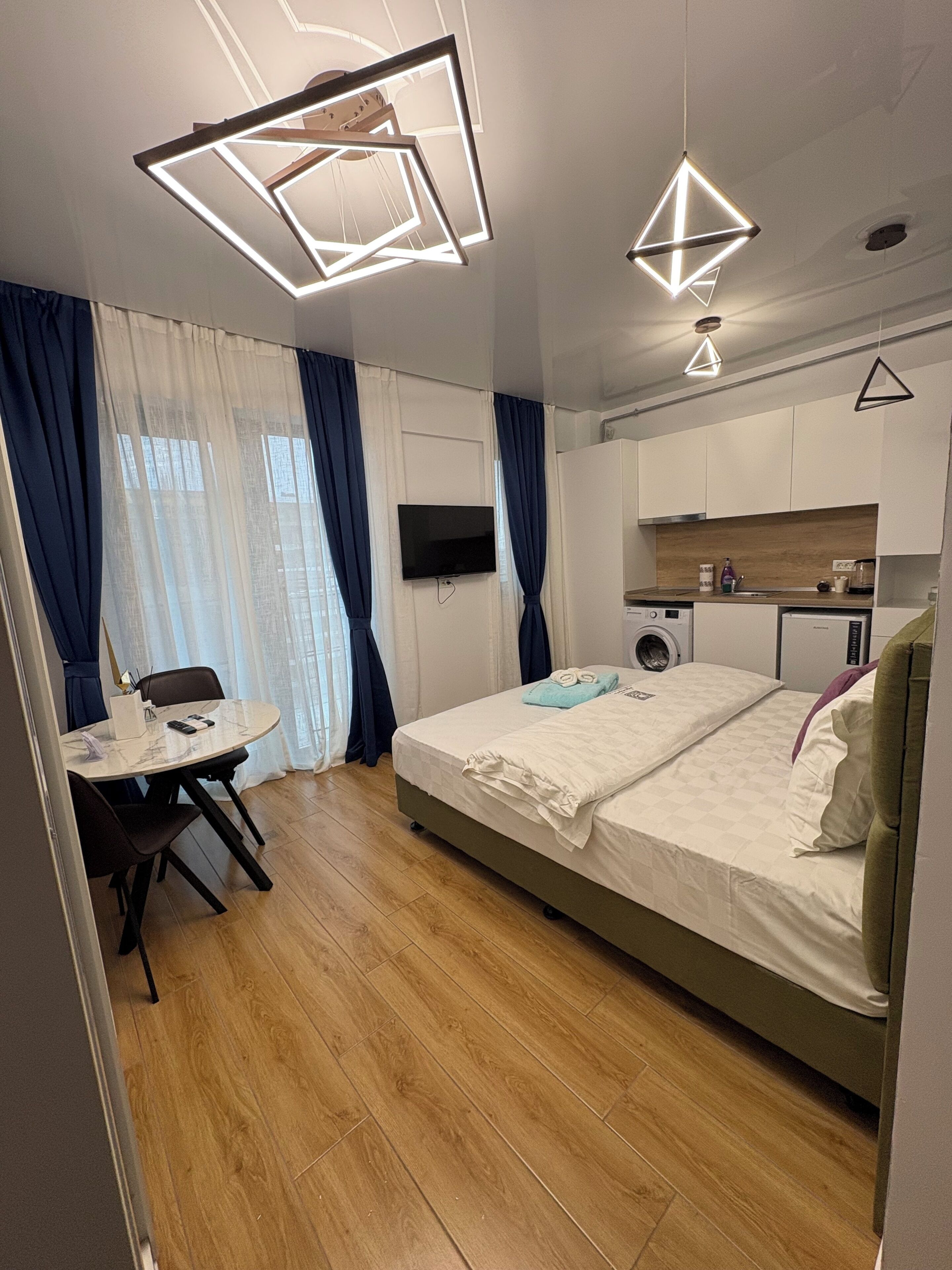 A modern hotel room with a bed, a small kitchen area, a dining table, and a TV.
