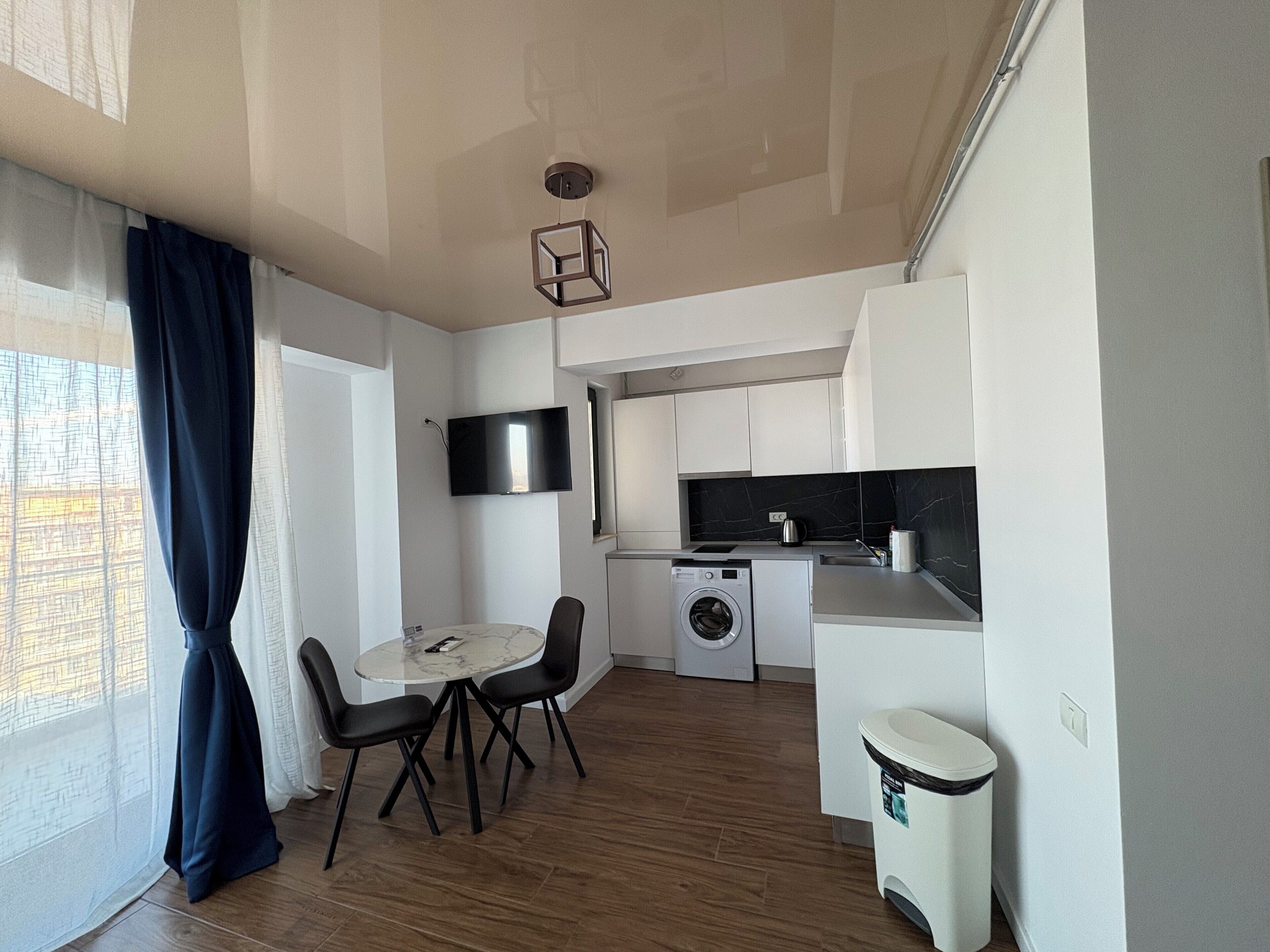 A compact kitchen with a washing machine, a dining area with a table and chairs, and a TV mounted on the wall.