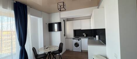 A compact kitchen with a washing machine, a dining area with a table and chairs, and a TV mounted on the wall.
