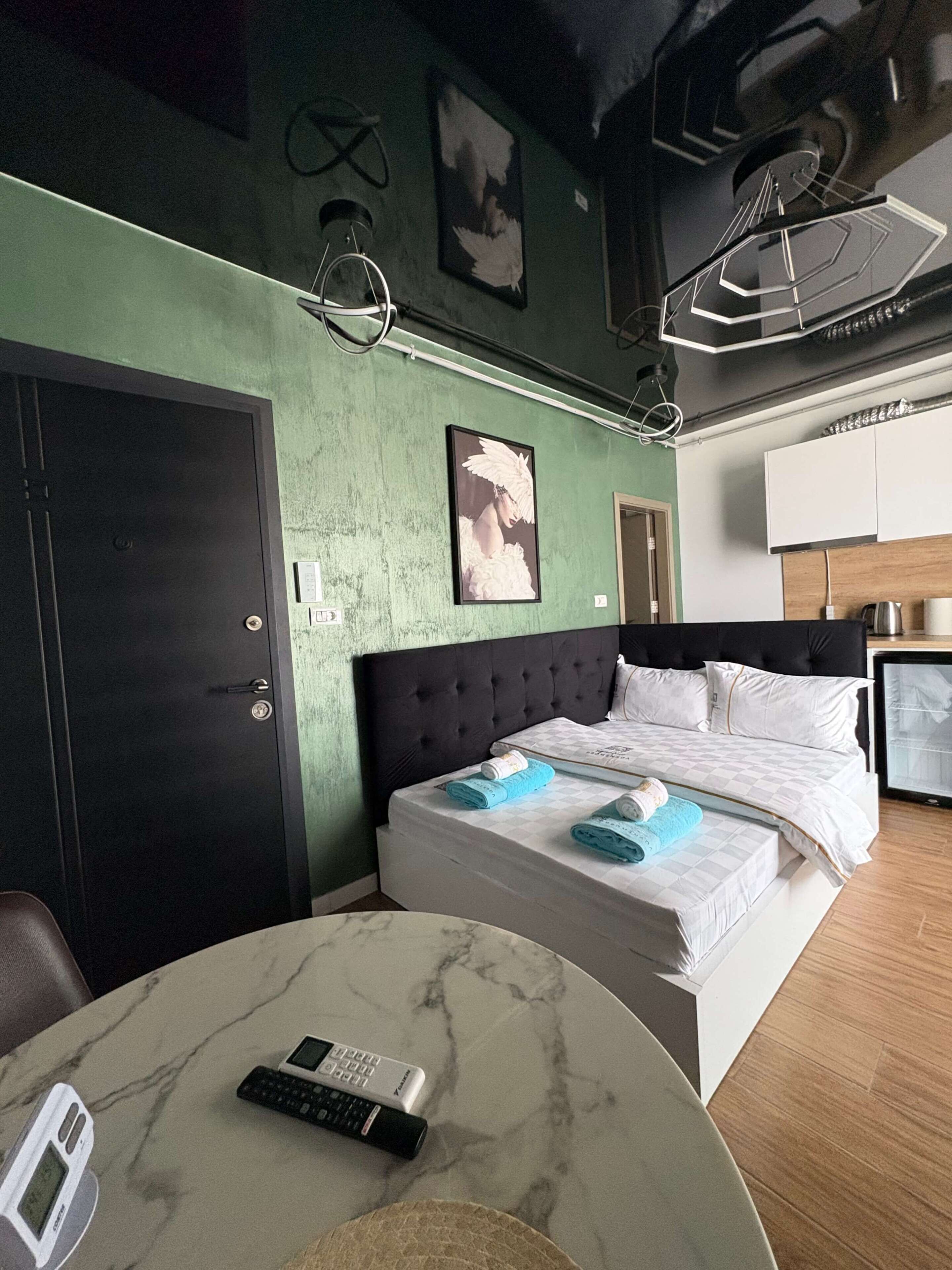 Hotellihuone, jossa on sänky, pyöreä pöytä, jossa on kaukosäädin, seinään kiinnitetty valaisin ja seinällä oleva kehystetty kuva.