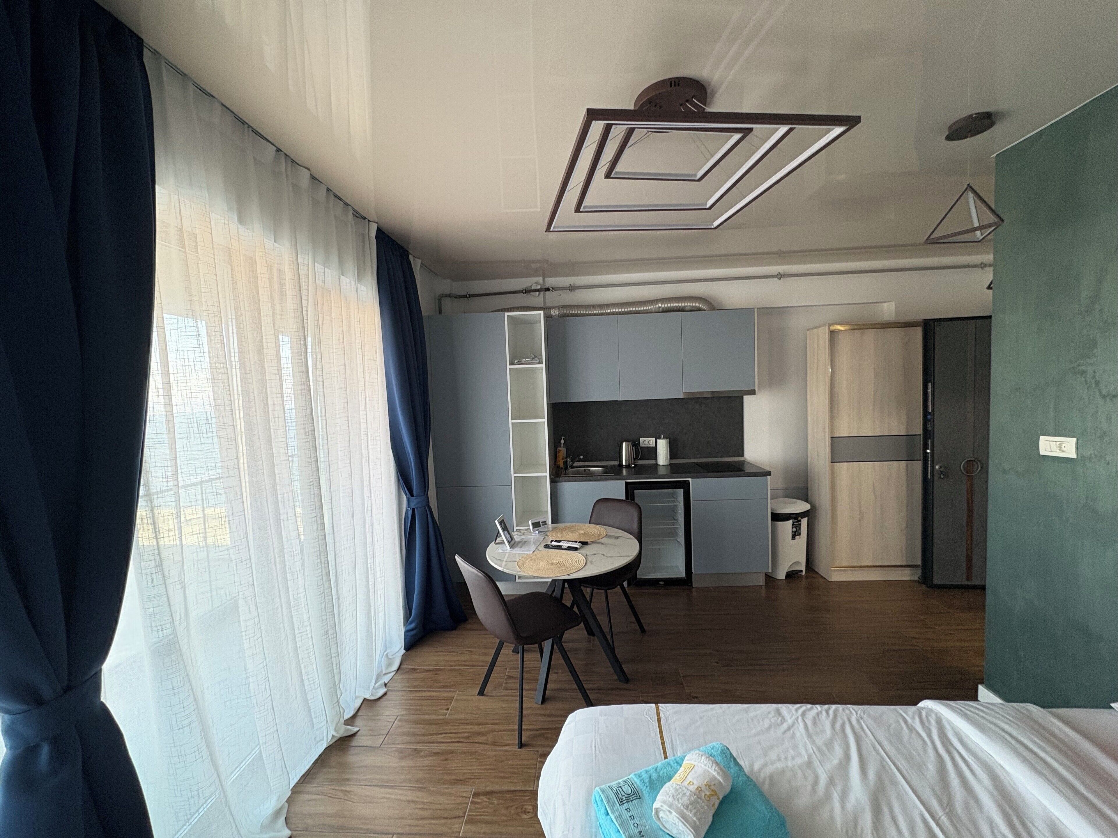A modern hotel room with a bed, a dining table with chairs, a kitchenette, and a large window with curtains.