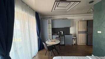 A modern hotel room with a bed, a dining table with chairs, a kitchenette, and a large window with curtains.