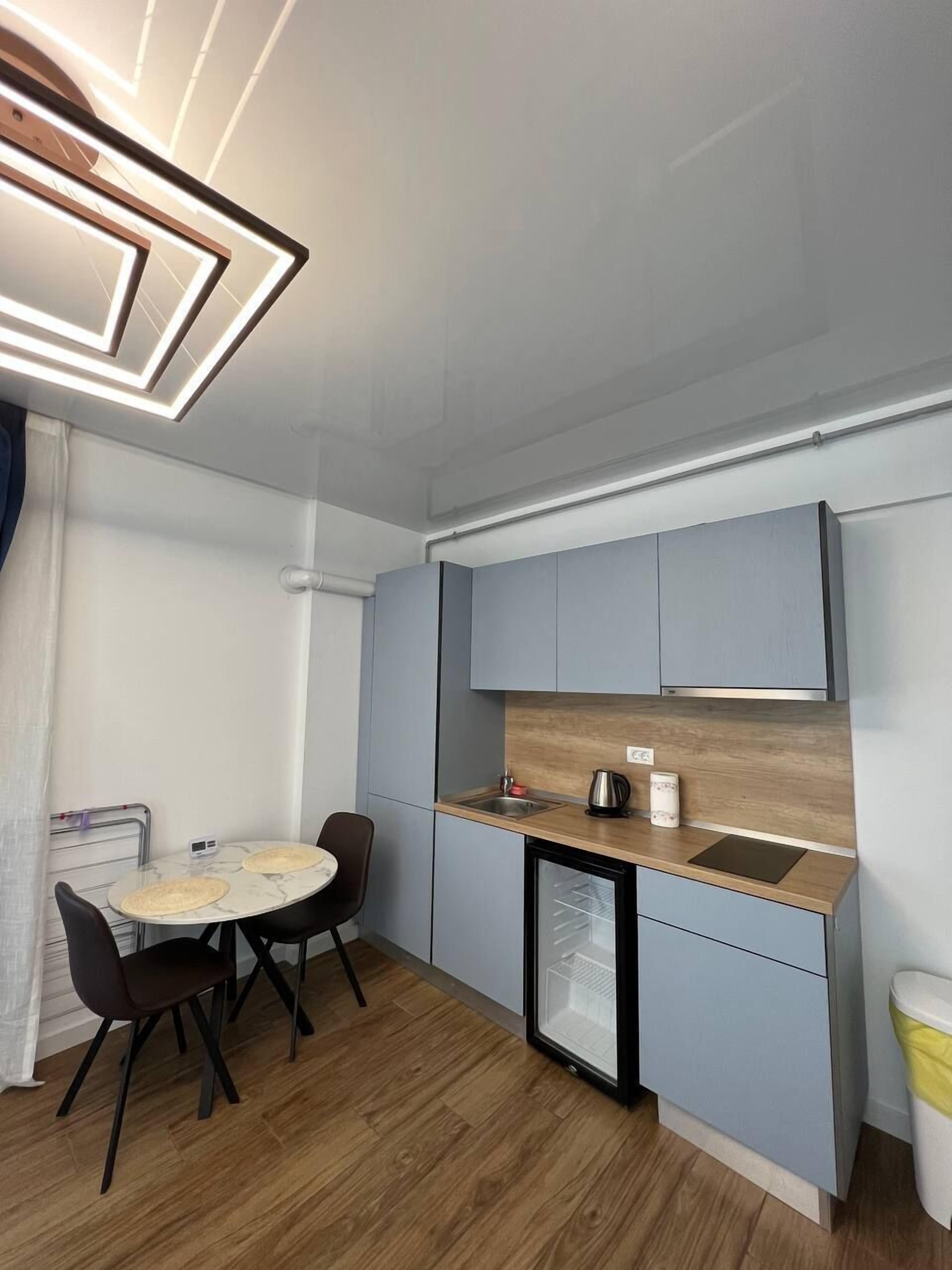 Une cuisine compacte équipée d’un réfrigérateur, d’un évier et d’un petit plan de travail. On y trouve une table à manger avec des chaises et un luminaire suspendu au plafond.
