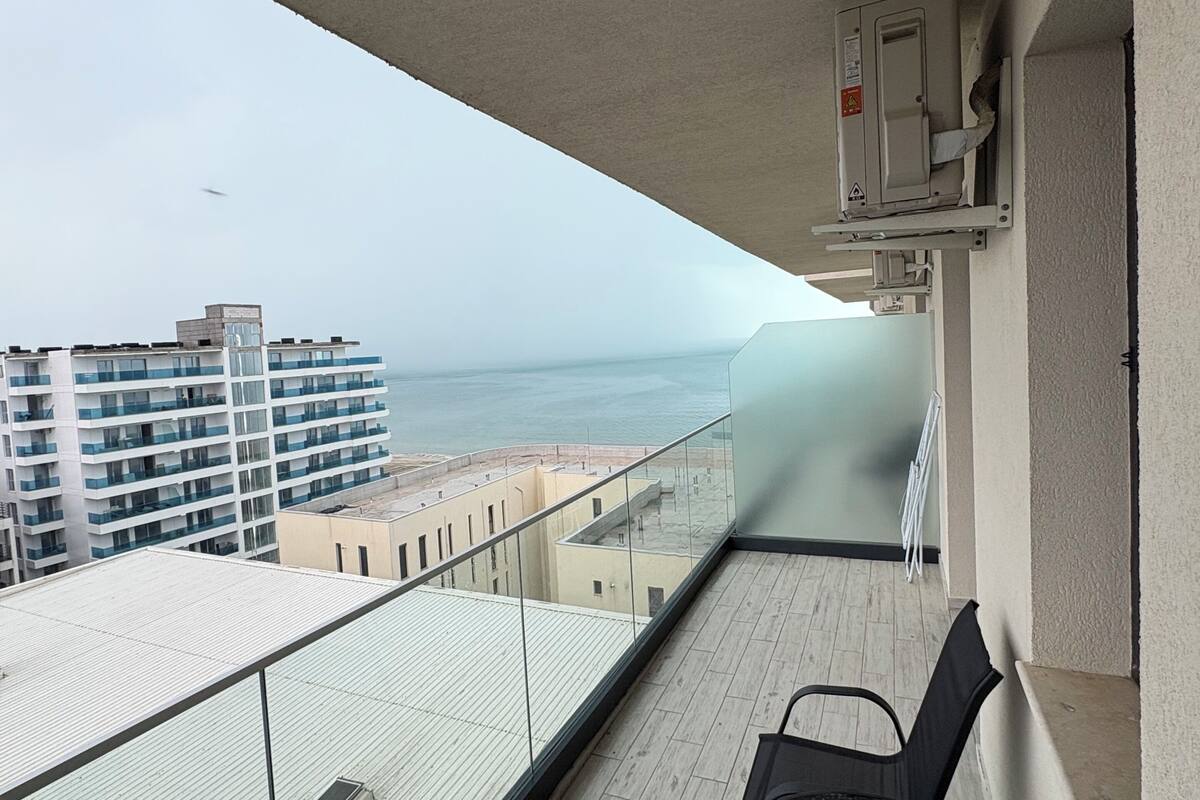 A balcony with a glass railing, a table, and chairs, overlooking a cityscape and the sea.