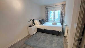 A small, neatly arranged bedroom with a bed, bedside tables, a lamp, and a view through a window.