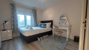 A bedroom with a bed, a desk with a mirror, a chair, a lamp, and a window with curtains.