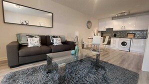 A modern living room with a glass coffee table, a sofa with decorative pillows, a dining area with a white chair, and a kitchen with white cabinets and a washing machine.