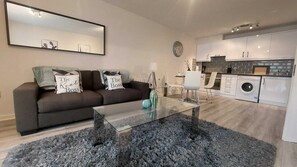 A modern living room with a glass coffee table, a sofa with decorative pillows, a dining area with a white chair, and a kitchen with white cabinets and a washing machine. - Modern 2BD @ Trinity College & Temple Bar (Dublin)