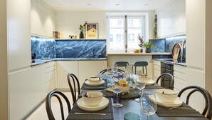 Ett modernt kök med vita skåp, en väggbeklädnad av marmor och en matplats med plats för fyra personer, bestående av ett bord, stolar och en fruktskål.
