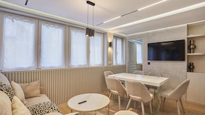 Een moderne kamer met een bank, een ronde tafel en een eettafel met stoelen. Er is een televisie aan de muur bevestigd en een raam met gordijnen.