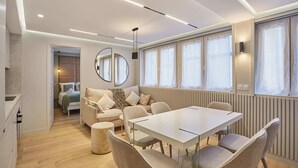 Een moderne woonkamer met een bank, een eettafel en stoelen. Er hangt een grote spiegel aan de muur en er is een raam met gordijnen.