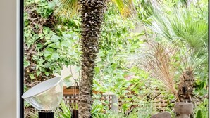 Una habitación con un amplio ventanal que da a un jardín exuberante, un mueble de madera oscura con objetos decorativos, un sillón blanco acolchado y un sofá de cuero.