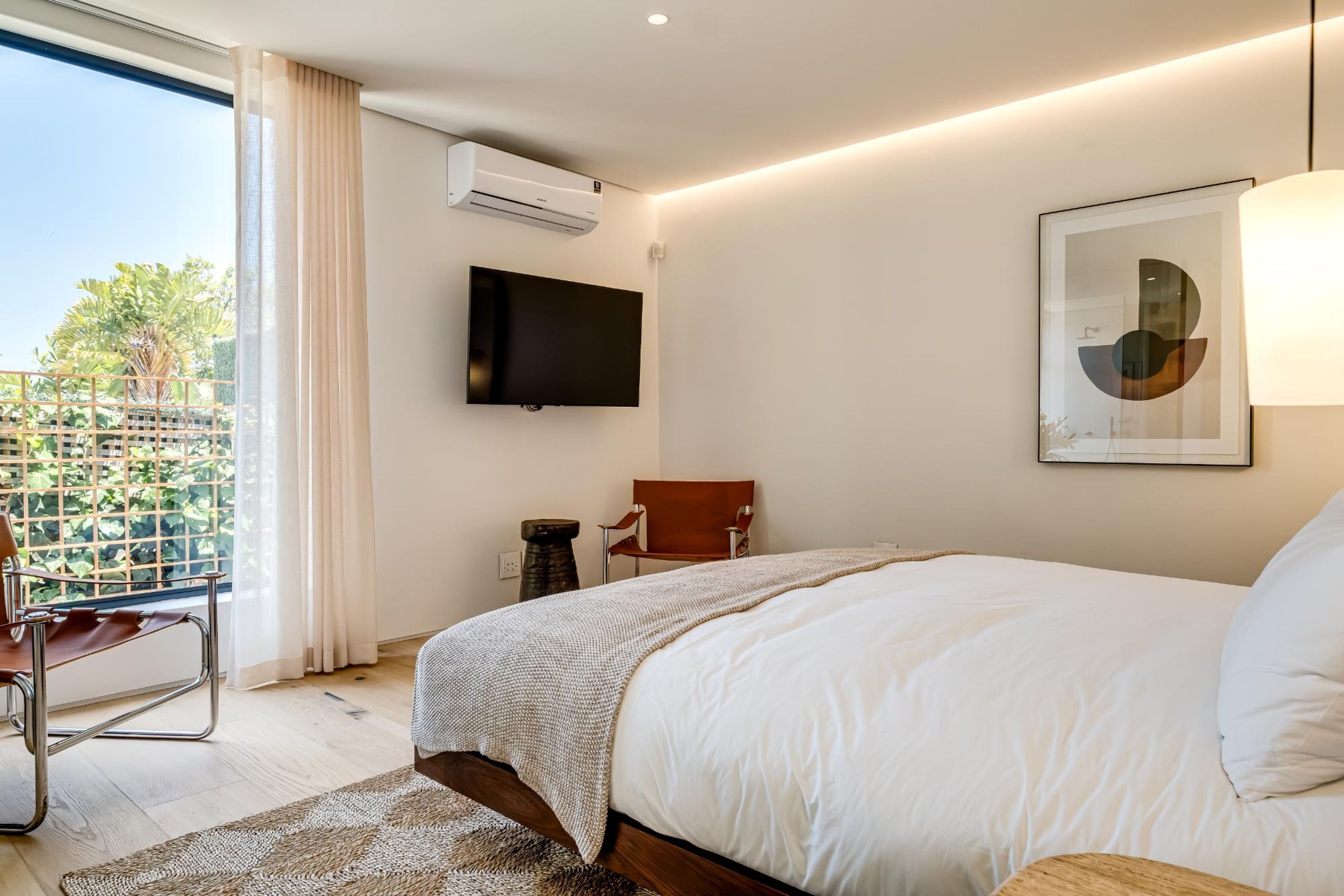 Ein modernes Schlafzimmer mit einem großen Bett, einem Wand-Fernseher, einem Sessel und einem Balkon mit Blick auf Grünflächen.