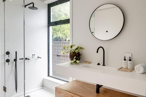 Une salle de bain moderne avec une douche en verre, un lavabo blanc avec un robinet noir, un miroir rond et une plante en pot.