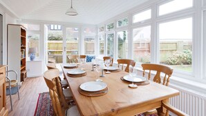 A dining area with a long wooden table set for six, surrounded by chairs, with large windows offering a view of the backyard. - Spacious 4-bed Seaside Retreat in Selsey (Chichester)