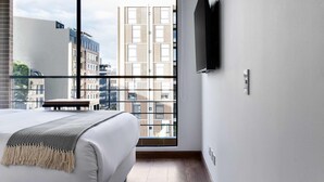 A modern hotel room with a large window, a bed with a white mattress and a patterned throw, a small table, and a mounted TV. - w Modern Loft w Brilliant Deco in Chapinero (Bogotá)