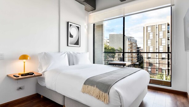 A modern bedroom with a large window, a bed with white bedding, a bedside table with a lamp, and a framed picture on the wall. - w Modern Loft w Brilliant Deco in Chapinero (Bogotá)