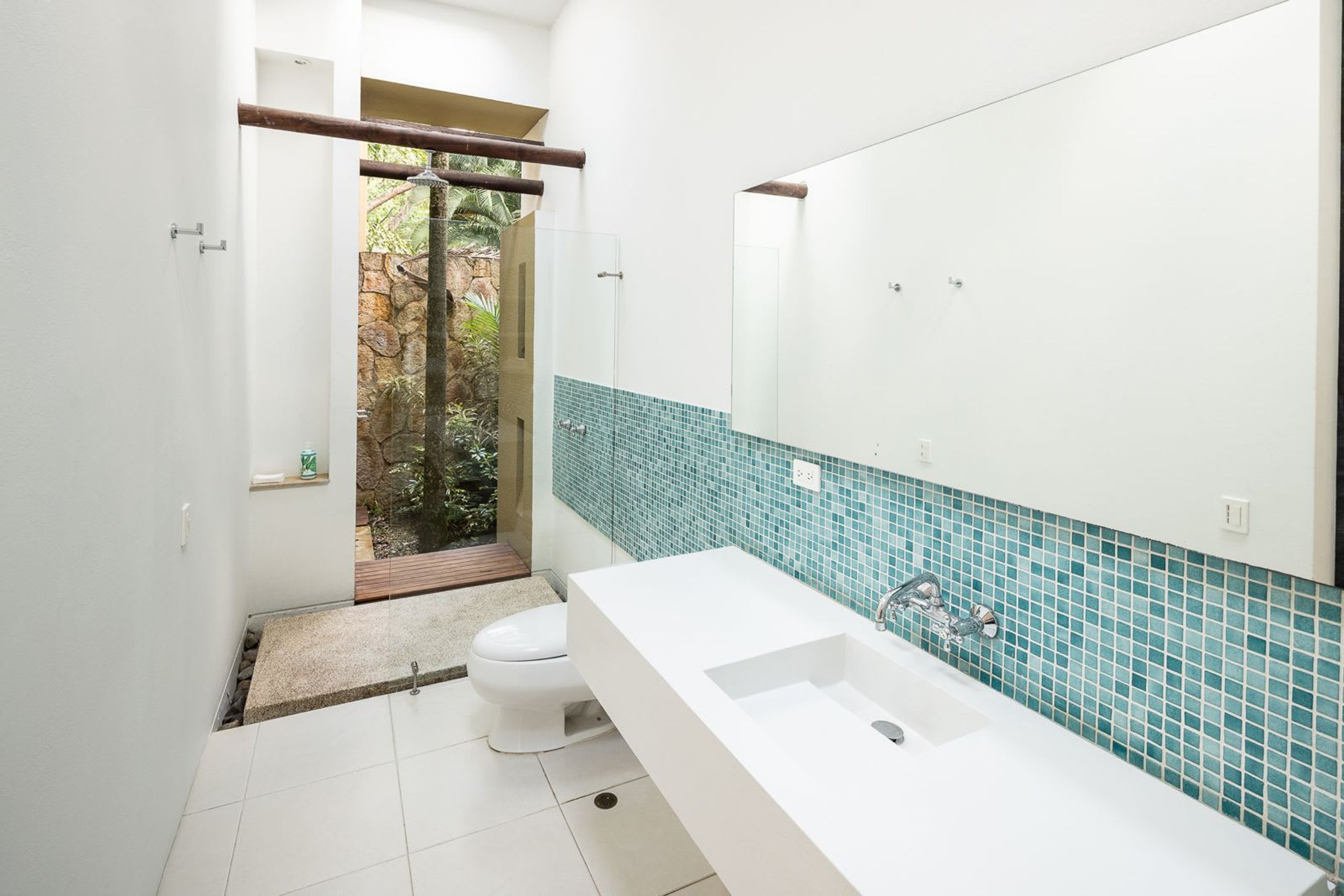 A modern bathroom with a white sink, a large mirror, and a view of a garden through a glass door.