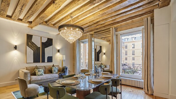 Un espace repas doté d’un plafond en bois, d’un lustre, d’une grande fenêtre avec des rideaux et d’un canapé agrémenté de coussins.