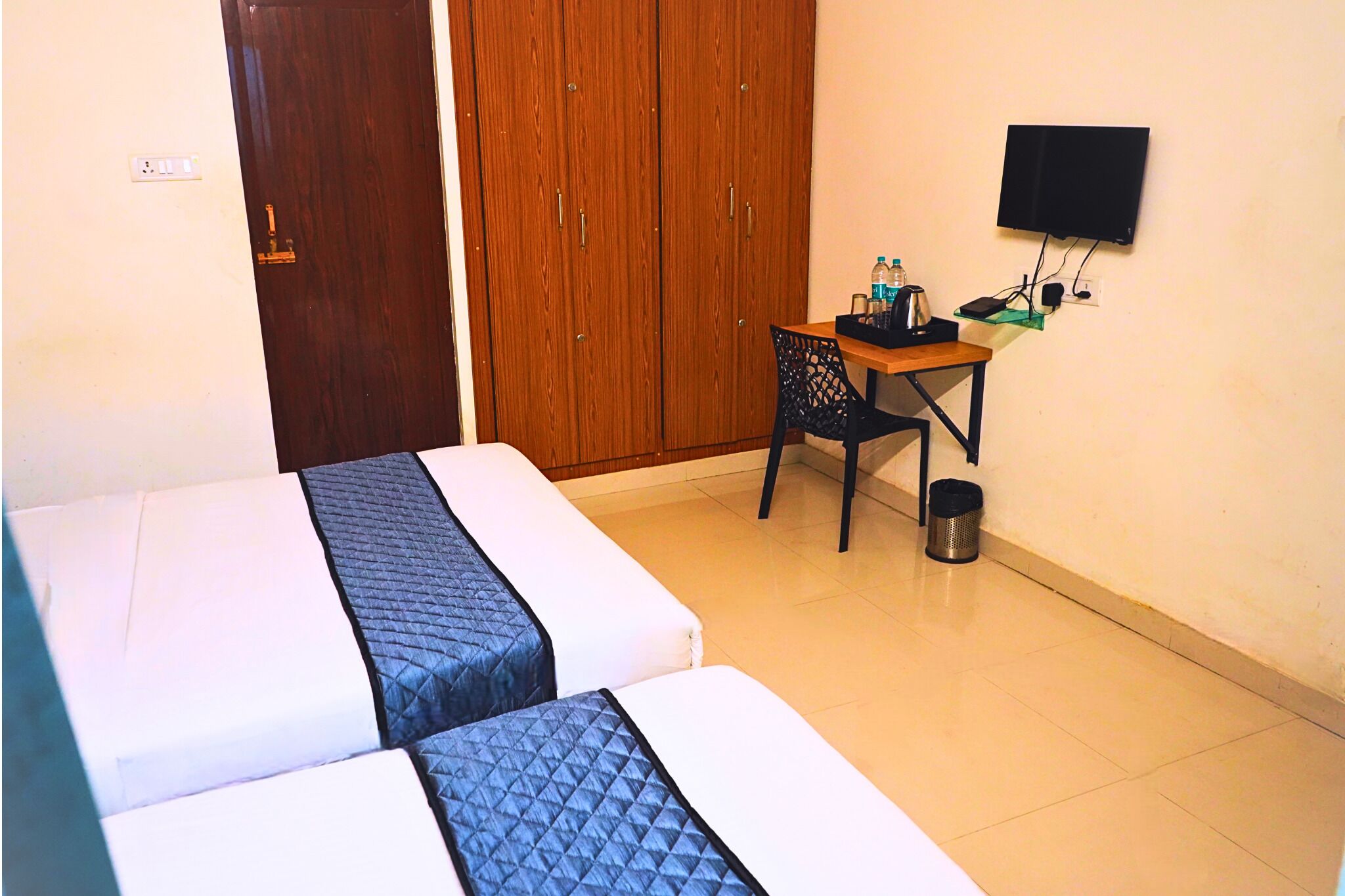 A hotel room with two beds, a wooden wardrobe, a desk with a chair, a television, and a trash bin.