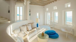 A spacious living room with a white sofa, a glass coffee table, and a blue ottoman. There are multiple windows providing views of the sea. - Elegant Villa With View (Mykonos)