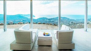 A covered outdoor seating area with white wicker furniture and a table, overlooking a mountainous landscape and the sea. - Elegant Villa With View (Mykonos)