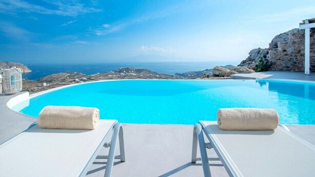 A clear blue infinity pool with two lounge chairs and towels, overlooking a coastal landscape and the sea. - Elegant Villa With View (Mykonos)