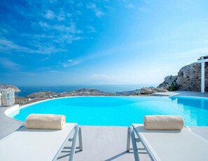 A clear blue infinity pool with two lounge chairs and towels, overlooking a coastal landscape and the sea.