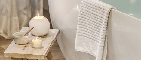 A spa bathtub with a white towel, a lit candle, and a small bowl with a spoon on a wooden stool.