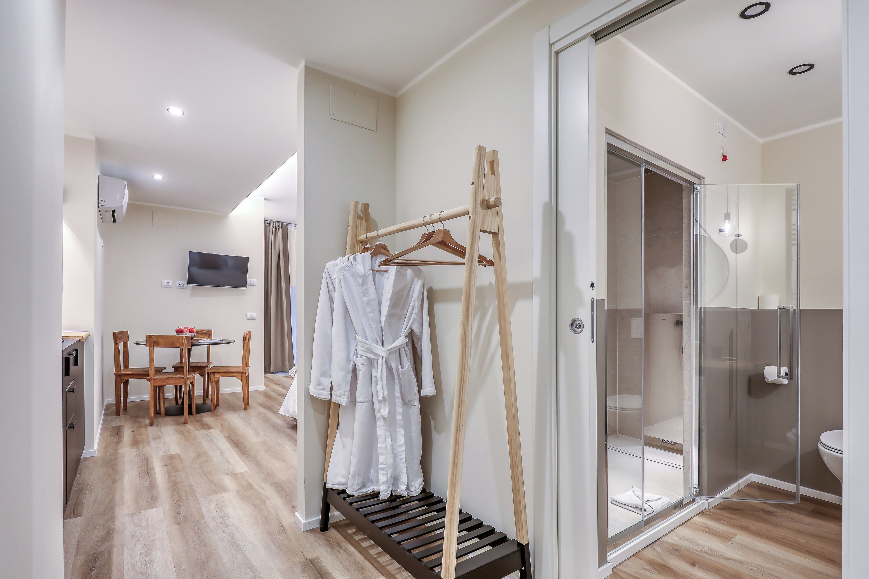 A hotel room with a wooden floor, a dining area with a table and chairs, a TV mounted on the wall, and a glass shower enclosure.