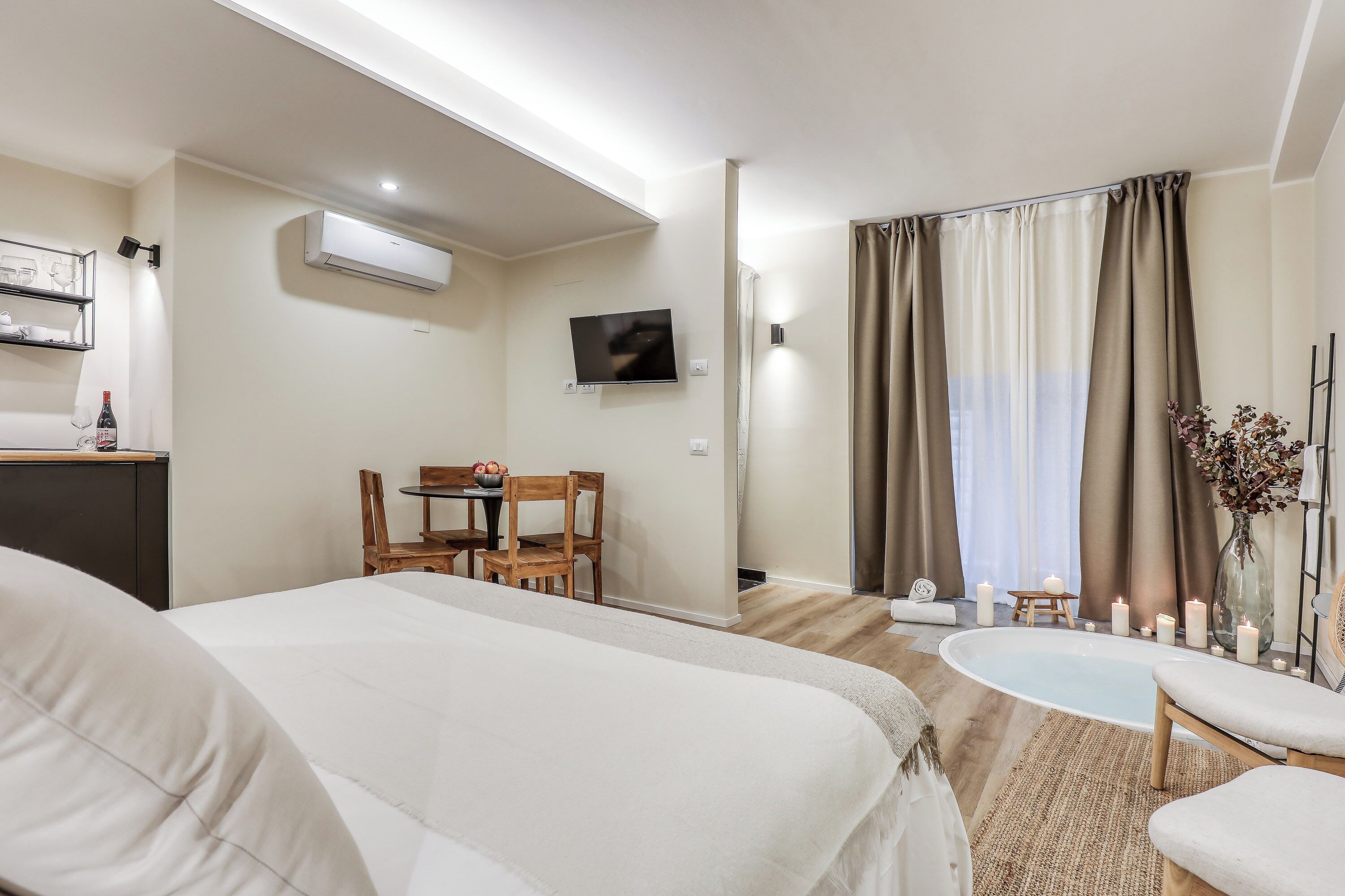 A hotel room with a large bed, a small dining table with chairs, a TV mounted on the wall, and a bathtub with candles.