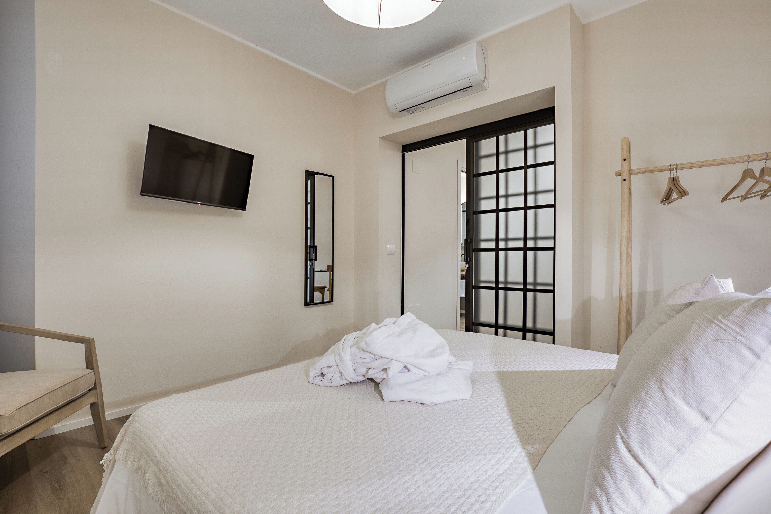 A hotel room with a bed, a TV mounted on the wall, a wooden chair, a robe on the bed, and a clothing rack with hangers.