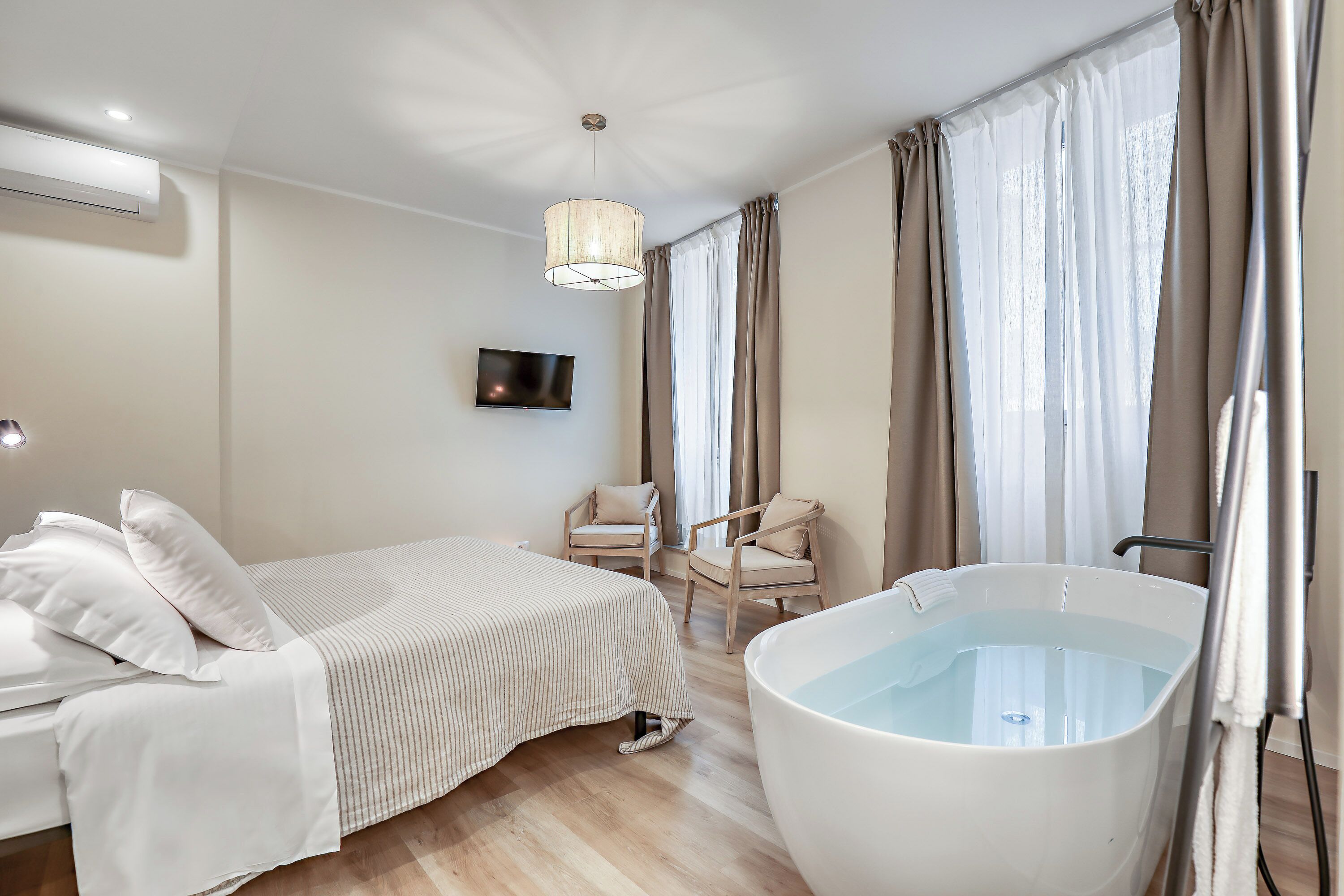 A bedroom with a bed, a bathtub, two chairs, and a TV on the wall.