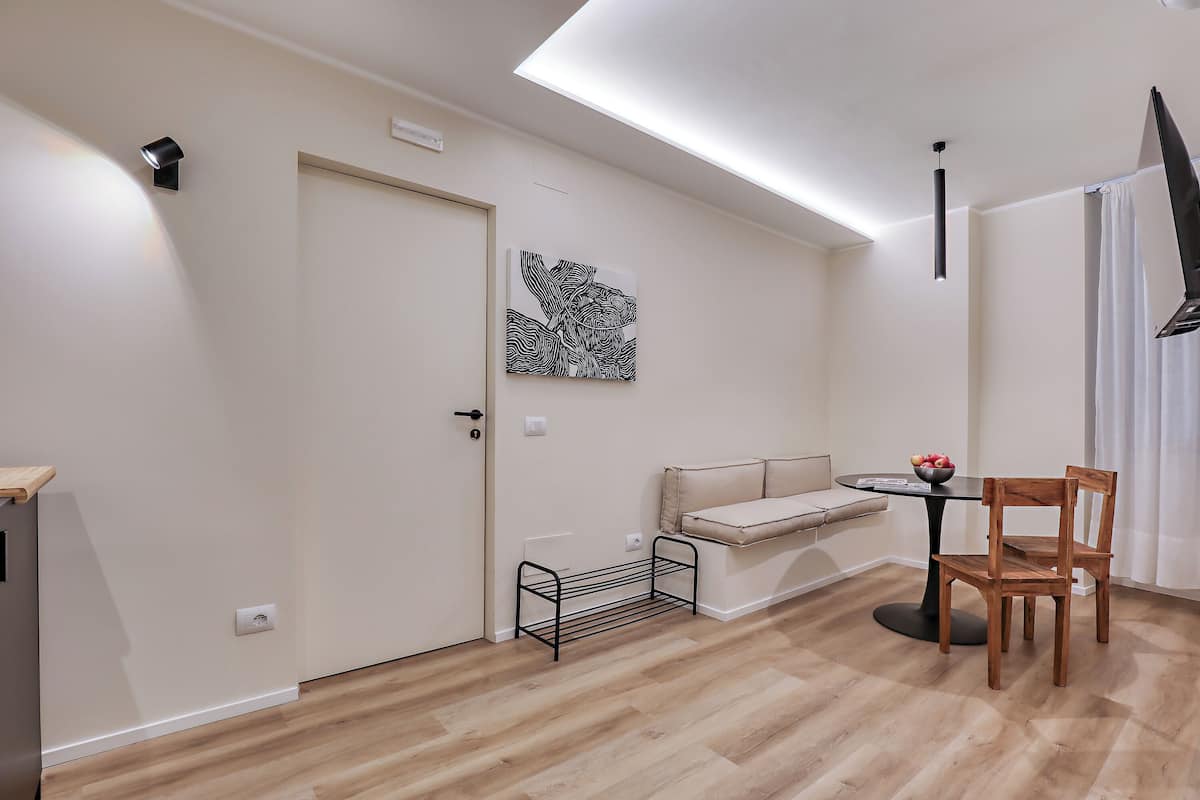 A modern living room with a wooden floor, a sofa, a dining table with chairs, a black metal shelf, and a wall-mounted artwork.