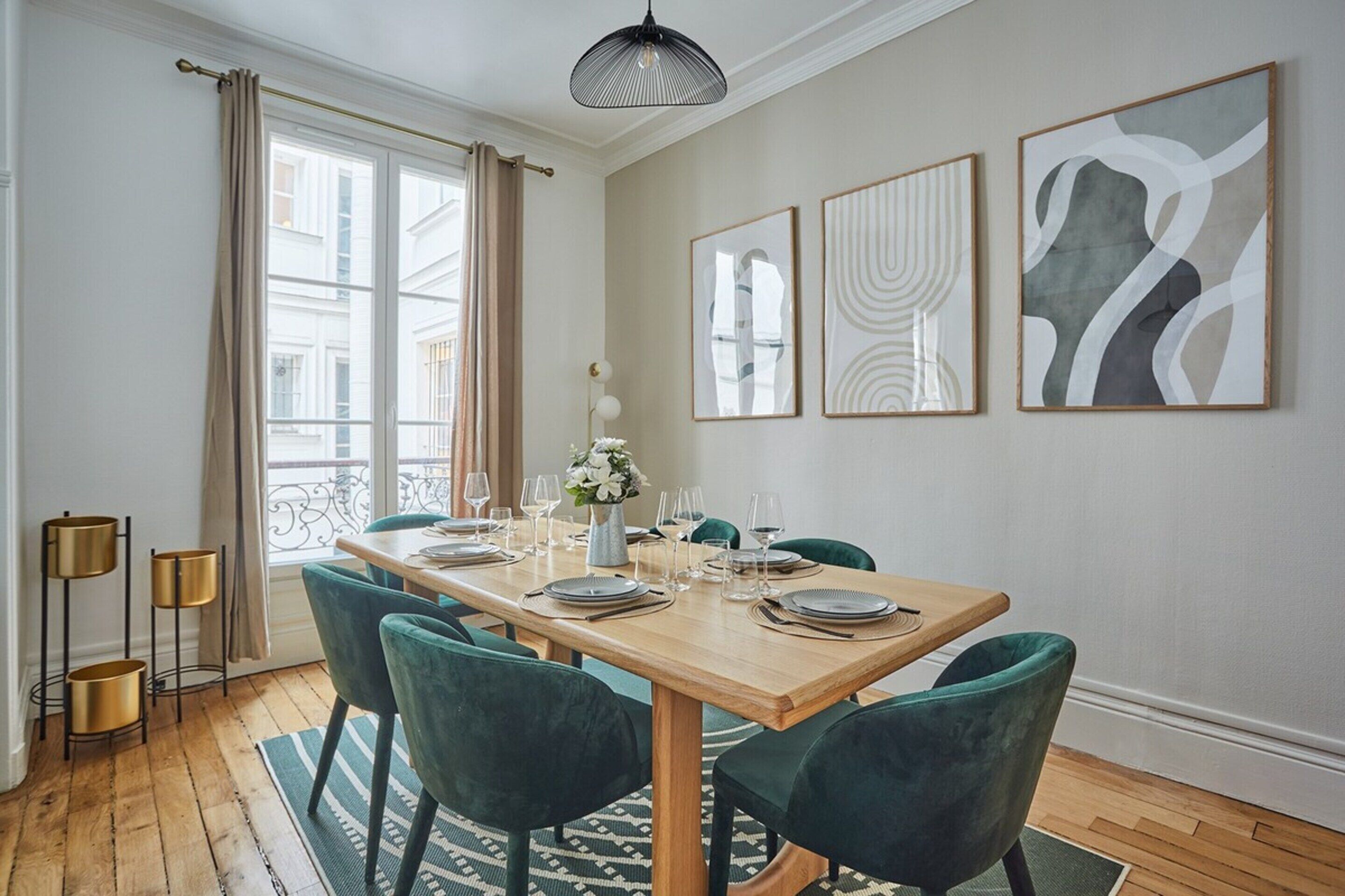 A dining area with a wooden table set for four, green upholstered chairs, a vase with flowers, and abstract wall art.