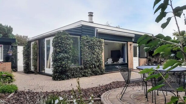 Una casa moderna con puerta de vidrio, sendero de ladrillo y jardín con plantas y una mesa.